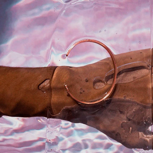 A Gold metal cuff bracelet with a small gap near one end, resting on a wet brown surface with pink and blue water ripples reflected in the background.